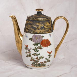 Pode incluir: Um bule de porcelana branca com alça e bico dourados. A seção superior apresenta uma faixa preta com ilustrações de paisagens em ouro. O corpo é decorado com motivos florais coloridos, incluindo flores roxas e laranjas e pequenas borboletas.