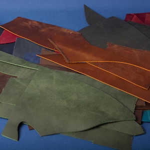 Può includere: Una collezione di pezzi di pelle di forma irregolare in vari colori, tra cui tonalità di marrone, verde, blu e bordeaux. I pezzi di pelle sono disposti su una superficie blu, mostrando le loro diverse trame e sfumature.