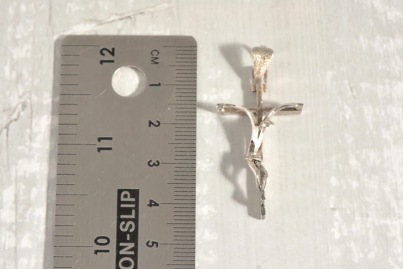 May include: A silver crucifix pendant. The pendant is approximately 5 cm in length and depicts Jesus on the cross in detail. The cross is positioned next to a ruler with cm measurements.
