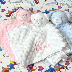 Peut inclure: Trois couvertures en peluche pour bébé avec des ours en peluche attachés. Les couvertures sont rose, blanche et bleue. La couverture blanche porte le nom "Maria" brodé.