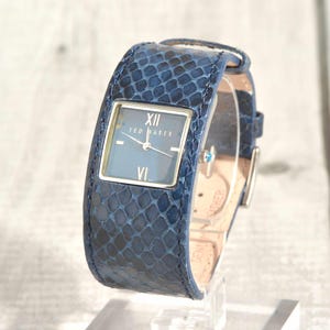 May include: A blue leather watch with a rectangular face. The watch face is blue with silver hands and Roman numerals. The watch strap has a snakeskin pattern. The watch is displayed on a clear stand.