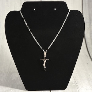 May include: A silver-coloured pendant necklace with a crucifix design. The pendant shows a figure on a cross. The necklace is displayed on a black jewellery stand against a light wood-grain background.
