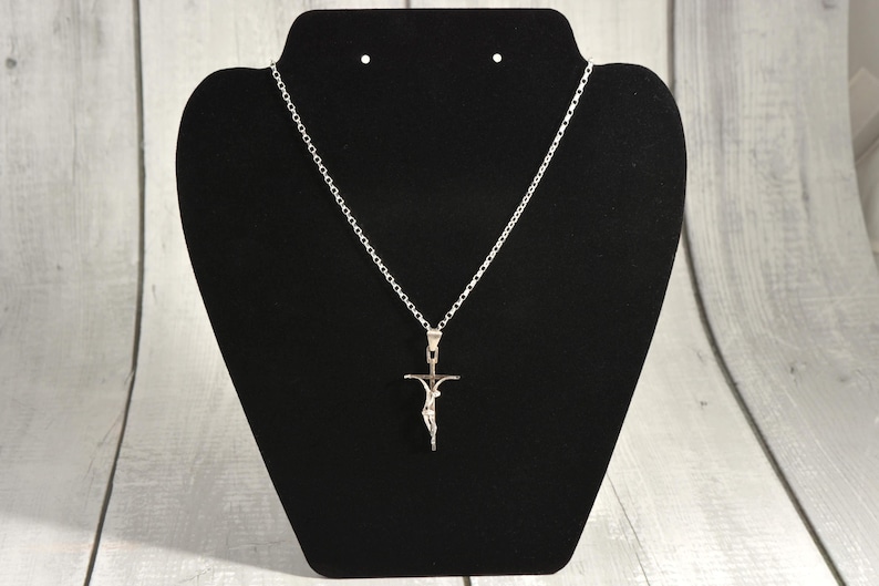 May include: A silver-coloured pendant necklace with a crucifix design. The pendant shows a figure on a cross. The necklace is displayed on a black jewellery stand against a light wood-grain background.