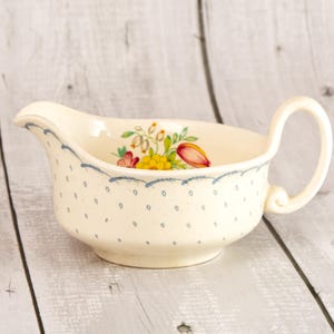 May include: A cream-colored ceramic gravy boat featuring a delicate blue dotted pattern across the exterior and a scalloped blue rim. The interior of the vessel is decorated with a colorful floral motif consisting of yellow, red, and green flowers.