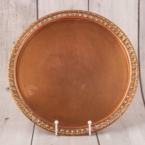 May include: A round, copper-coloured tray with an ornate border. The border has a repeating pattern of small, raised details in a gold tone. The tray is displayed on a white stand against a light wood-grain background.