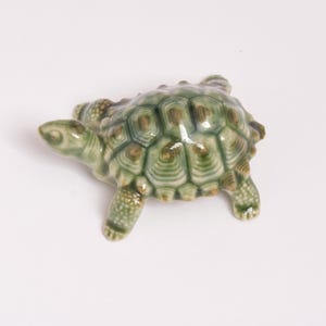 May include: A small ceramic turtle figurine featuring a glossy finish with shades of sage green and brown. The shell displays a detailed geometric pattern of raised hexagonal plates, while the head and legs show a textured, speckled surface.