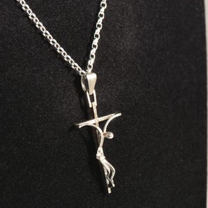 May include: A silver pendant necklace with a crucifix design. The pendant shows a stylised figure on a cross, suspended from a delicate chain. The necklace is displayed against a dark background, highlighting the intricate details of the jewellery.