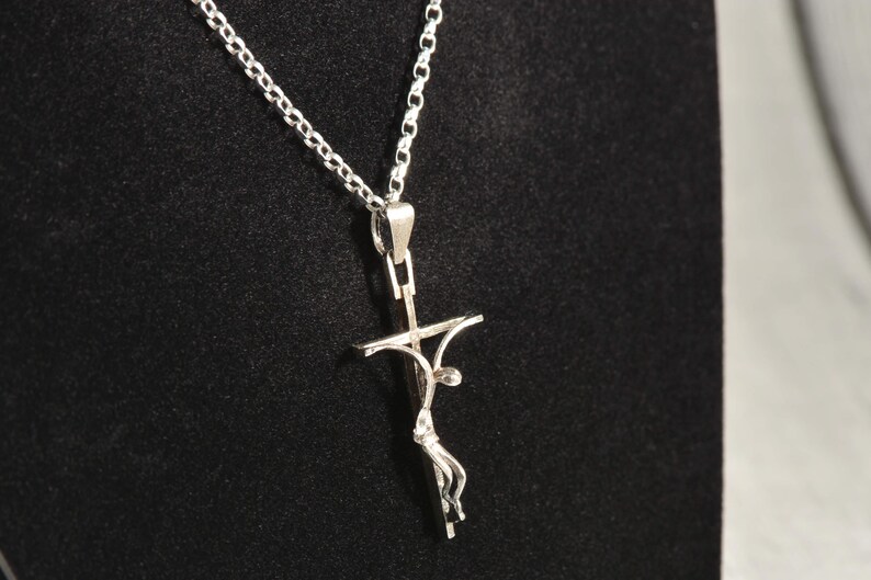 May include: A silver pendant necklace with a crucifix design. The pendant shows a stylised figure on a cross, suspended from a delicate chain. The necklace is displayed against a dark background, highlighting the intricate details of the jewellery.
