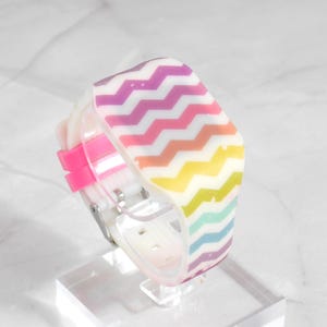 May include: A digital watch with a white strap and a pink clasp. The watch face has a multi-coloured chevron pattern in pink, purple, orange, yellow, green, and blue. The watch is displayed on a clear stand.