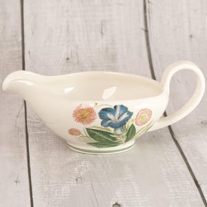 May include: A cream-coloured ceramic gravy boat featuring a botanical illustration of a large blue flower flanked by smaller pink and orange blossoms with green leaves. The vessel has a curved handle and a pouring spout on the opposite side.