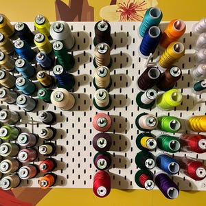 May include: A white pegboard filled with colorful spools of thread. The thread colors include green, yellow, blue, red, pink, and brown. The spools are arranged in neat rows, creating a visually appealing display for sewing or crafting.