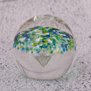 Könnte beinhalten: Ein klarer Glas-Briefbeschwerer mit farbenfrohem Design. Das Innere zeigt eine Ansammlung blauer, grüner und weißer Punkte, die einen gesprenkelten Effekt erzeugen. Der Briefbeschwerer hat eine abgerundete Form mit spitzer Basis.