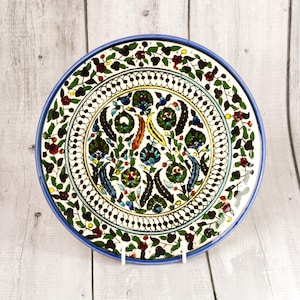 May include: A round ceramic plate featuring a hand-painted floral pattern with dark green, red, and blue motifs on a cream background. The border is decorated with a repeating vine design, and the entire piece is framed by a solid blue rim.