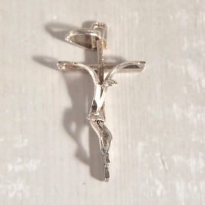 May include: A silver crucifix pendant with a detailed depiction of Jesus. The pendant has a textured surface and is suspended from a small loop. The cross arms are slightly curved.