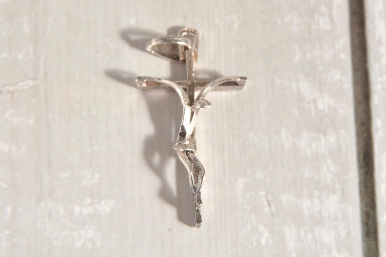 May include: A silver crucifix pendant with a detailed depiction of Jesus. The pendant has a textured surface and is suspended from a small loop. The cross arms are slightly curved.