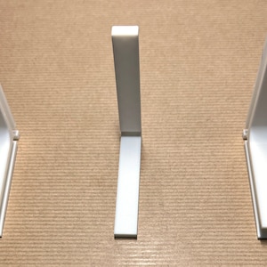 May include: Three white plastic corner brackets, each with a 90-degree angle, are arranged in a row on a brown surface.
