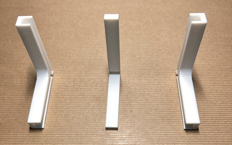 May include: Three white plastic corner brackets, each with a 90-degree angle, are arranged in a row on a brown surface.