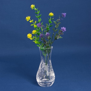 Può includere: Un vaso in vetro trasparente con bordo svasato e collo affusolato, caratterizzato da un motivo floreale inciso sul corpo. Il vaso contiene diversi steli di fiori artificiali con piccoli fiori gialli e viola e foglie verdi.