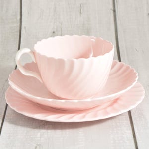 May include: A pale pink teacup, saucer, and side plate set. The teacup has a curved handle and a fluted rim. The saucer and side plate also have fluted edges. The set is made of ceramic and is on a light wood surface.