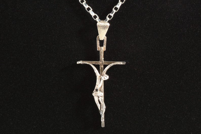 May include: A silver crucifix pendant necklace. The pendant shows a detailed figure of Jesus Christ on the cross. The necklace has a simple chain and is set against a black background.