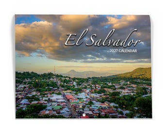 May include: A wall calendar featuring a panoramic landscape photograph of a town in El Salvador at sunset. The sky is filled with golden and blue clouds above a dense collection of buildings and green hills. The text reads El Salvador 2027 CALENDAR.