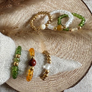 Könnte beinhalten: Satz von fünf Perlenringen mit goldenen Akzenten. Die Ringe sind aus grünen, weißen und orangefarbenen Perlen gefertigt. Ein Ring hat eine rote Steinperle, ein anderer eine weiße Steinperle und ein anderer eine gelbe Steinperle.