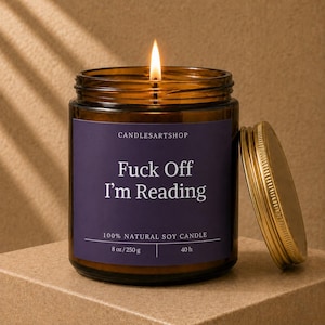 May include: An amber glass jar candle with a lit wick and a gold metal lid. The dark purple label reads CANDLESARTSHOP, Fuck Off I'm Reading, 100% NATURAL SOY CANDLE, 8 oz / 250 g, and 40 h. The candle is filled with wax and emits a bright flame.
