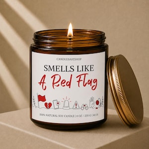 Peut inclure: Une bougie en pot de verre brun avec une étiquette blanche portant le texte "SMELLS LIKE A Red Flag" en noir et rouge. L'étiquette présente des icônes rouges et noires représentant un drapeau, un cœur brisé, un téléphone, une sirène et plus.