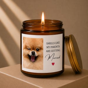 Peut inclure: Une bougie allumée dans un pot en verre ambré avec un couvercle en métal. L'étiquette blanche présente une photo d'un chien Poméranien qui fait un clin d'œil et le texte noir : SMELLS LIKE MY PARENTS ARE GETTING Married, avec un petit cœur rouge.