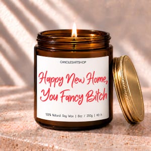 May include: A lit candle in an amber glass jar with a gold lid. The white label features the text CANDLESARTSHOP, Happy New Home, You Fancy Bitch, 100% Natural Soy Wax, 8oz, 250g, and 40 h in black and red script and sans-serif fonts.