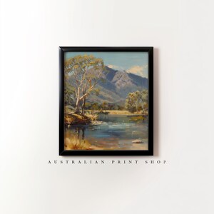 May include: A framed painting of a landscape scene with a lake, mountains, and trees. The sky is blue with white clouds. The lake is a light blue color and reflects the sky. The mountains are a dark gray color. The trees are a dark green color. The painting is in a black frame.
