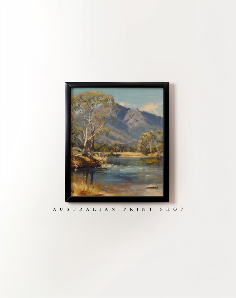 May include: A framed painting of a landscape scene with a lake, mountains, and trees. The sky is blue with white clouds. The lake is a light blue color and reflects the sky. The mountains are a dark gray color. The trees are a dark green color. The painting is in a black frame.