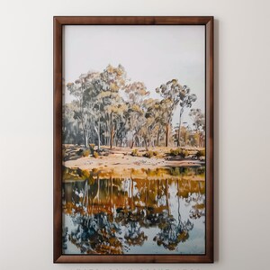 May include: A framed print of a landscape painting featuring a reflection of trees in a still body of water. The trees are a mix of green and brown, and the water is a deep blue. The frame is a dark wood.