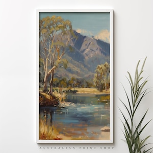 May include: A framed painting of a landscape scene with a river, trees, and mountains in the background. The painting is in a white frame and has a blue and green color scheme. The text "AUSTRALIAN PRINT SHOP" is at the bottom of the frame.