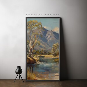May include: A framed painting of a landscape scene with a river, trees, and mountains in the background. The painting is in a black frame and is hanging on a white wall. The text "AUSTRALIAN PRINT SHOP" is printed above the painting.