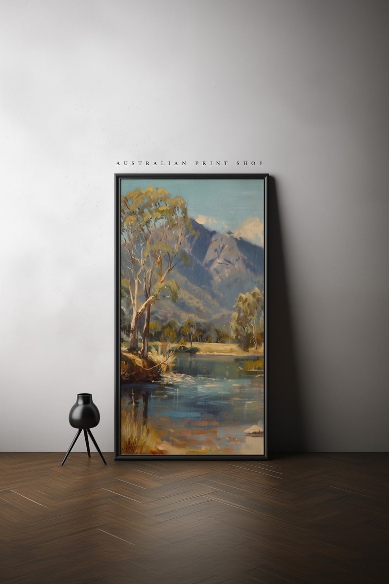 May include: A framed painting of a landscape scene with a river, trees, and mountains in the background. The painting is in a black frame and is hanging on a white wall. The text "AUSTRALIAN PRINT SHOP" is printed above the painting.
