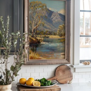 May include: A framed landscape painting of a river and mountains hangs on a white tiled wall above a kitchen counter. The painting is in a rustic wooden frame. On the counter, there is a wooden cutting board with a bowl of lemons and a bowl of green vegetables.
