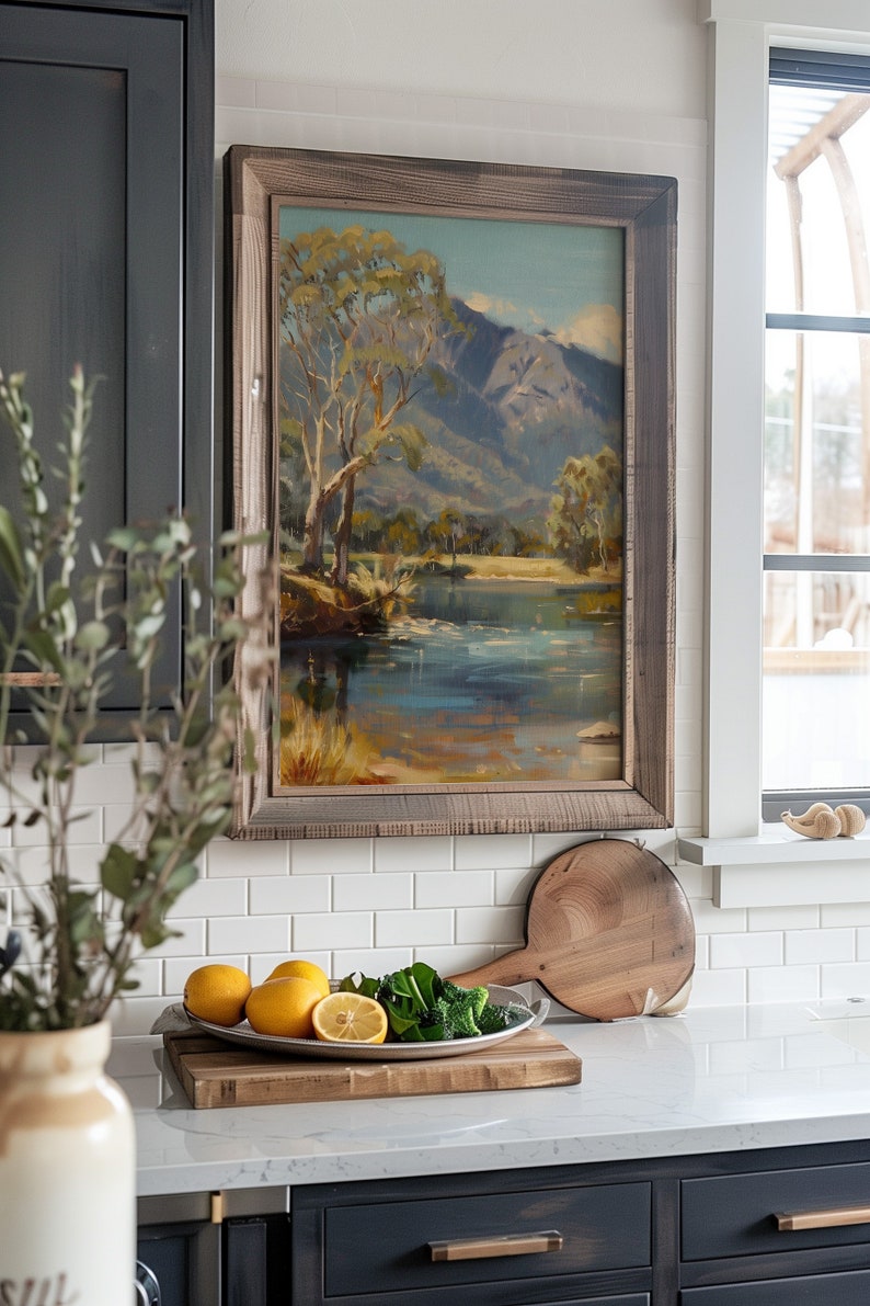 May include: A framed landscape painting of a river and mountains hangs on a white tiled wall above a kitchen counter. The painting is in a rustic wooden frame. On the counter, there is a wooden cutting board with a bowl of lemons and a bowl of green vegetables.