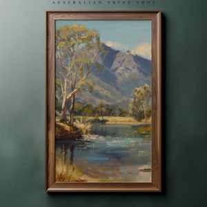 May include: A framed painting of a landscape scene with a river, trees, and mountains in the background. The painting is in a wooden frame and has a rustic feel.