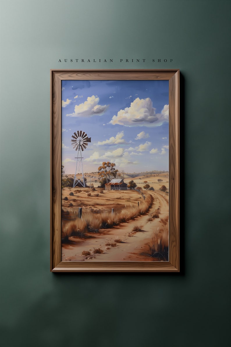 May include: A framed painting of a rural Australian landscape with a windmill, a farmhouse, and a dirt road winding through the dry, brown fields under a blue sky with white clouds.