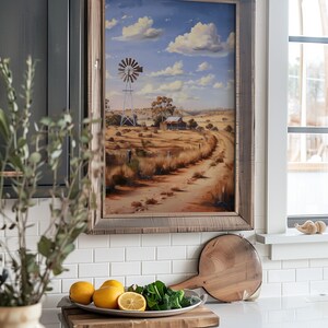 May include: A framed painting of a rural landscape with a windmill, a farmhouse, and a dirt road leading into the distance. The sky is blue with white clouds. The painting is hanging on a white tiled wall in a kitchen.