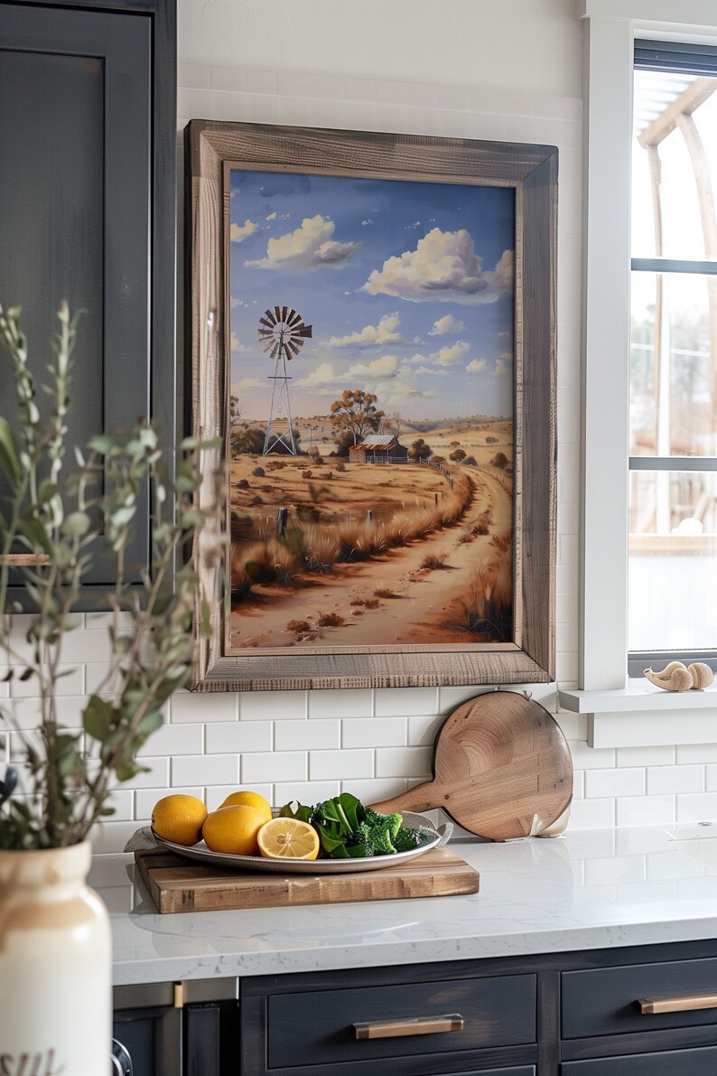 May include: A framed painting of a rural landscape with a windmill, a farmhouse, and a dirt road leading into the distance. The sky is blue with white clouds. The painting is hanging on a white tiled wall in a kitchen.