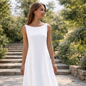 Puede incluir: Vestido blanco sin mangas, con cuello barco y falda en forma de A. El vestido llega a la rodilla y está hecho de una tela ligera. La persona lleva sandalias marrones. El fondo muestra un camino de piedra y vegetación.