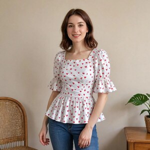 May include: A white top patterned with red hearts, featuring a square neckline and short, flared sleeves. The top has a peplum waist. The model is wearing blue jeans.