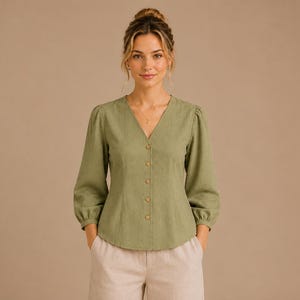 May include: A sage green button-down blouse featuring a V-neckline, long sleeves with gathered cuffs, and a fitted bodice. The garment is styled with light beige trousers. The blouse has a textured fabric finish and five visible round buttons.