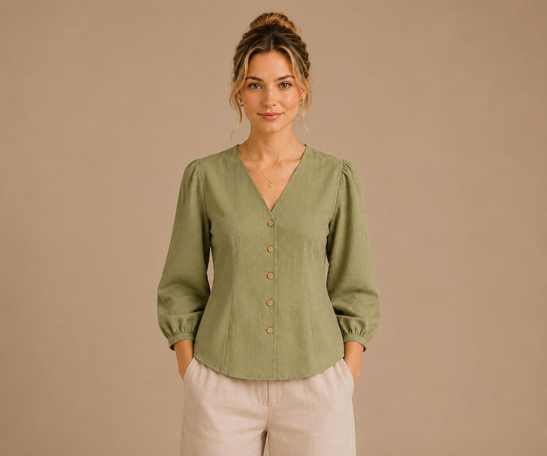 May include: A sage green button-down blouse featuring a V-neckline, long sleeves with gathered cuffs, and a fitted bodice. The garment is styled with light beige trousers. The blouse has a textured fabric finish and five visible round buttons.