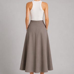 May include: A white sleeveless racerback tank top paired with a solid taupe midi skirt. The skirt features a high-waisted design, a flared A-line silhouette, and side pockets. The ensemble is completed with beige heeled sandals.