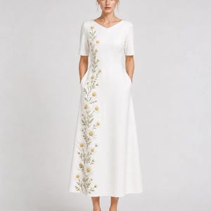May include: A white midi-length dress featuring a V-neckline, short sleeves, and side pockets. The garment is adorned with a vertical floral embroidery pattern of white daisies with yellow centers and green stems running down the right side.