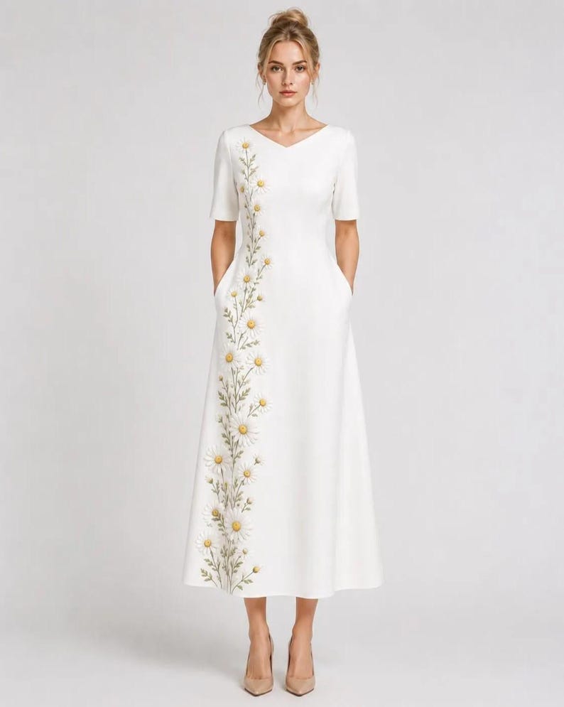 May include: A white midi-length dress featuring a V-neckline, short sleeves, and side pockets. The garment is adorned with a vertical floral embroidery pattern of white daisies with yellow centers and green stems running down the right side.