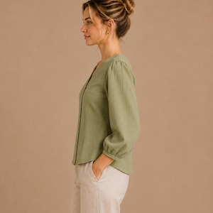 May include: A sage green long-sleeve button-down shirt with a V-neckline and gathered cuffs, paired with light beige trousers. The shirt features a straight hemline and a simple, clean design, showcasing a soft, textured fabric appearance.